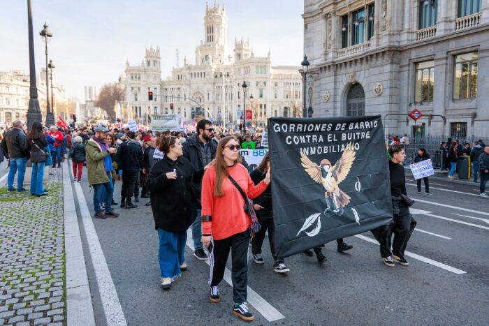 Manifestantes marchan pola rúa de Alcalá durante a «Manifestación por unha vivenda digna» en Madrid o 9 de febreiro de 2025, co Palacio de Cibeles ao fondo. A súa pancarta negra reza «Gorriones de barrio contra o voitre inmobiliario», unha metáfora poética e poderosa profundamente arraigada nos movementos sociais urbanos. O termo «gorriones» simboliza aos residentes locais, pequenos, resistentes e profundamente conectados co seu barrio, que loitan contra os «voitres inmobiliarios», grandes fondos de investimento depredadores que se aproveitan das comunidades urbanas.